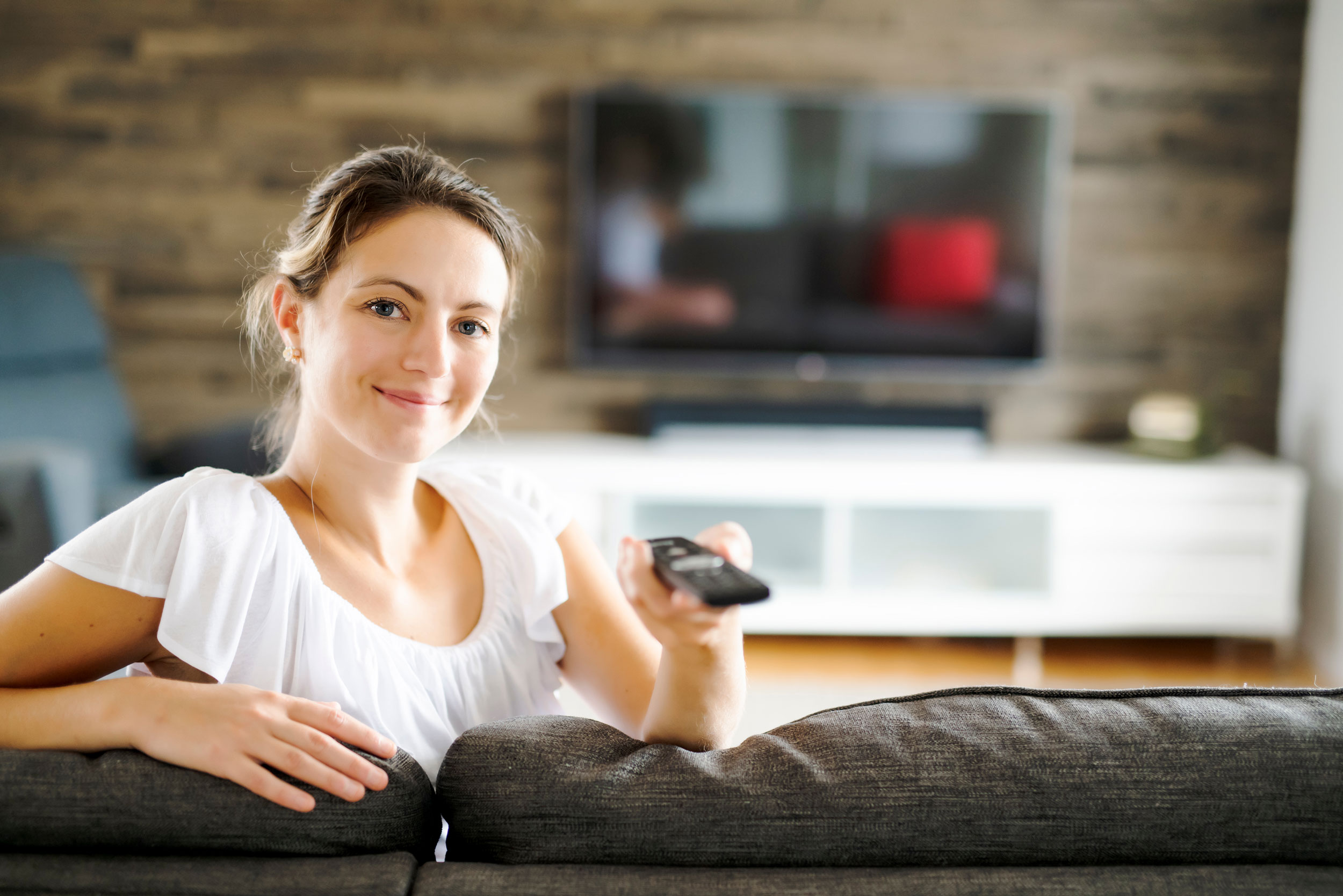 Frau mit Fernbedienung auf einem Sofa sitzend, im Hintergrund ein unscharfer Fernseher